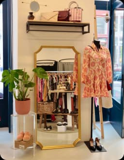 Entrée de la boutique avec un miroir doré, un mannequin en robe fleurie orange et une étagère de sacs à main