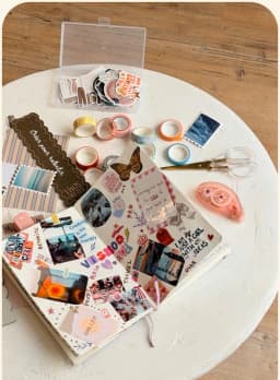 Table ronde blanche couverte de matériel de scrapbooking : carnet illustré de stickers, rubans washi, ciseaux et tampons