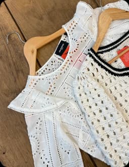 Deux hauts blancs sur cintres posés sur une table en bois, l'un en broderie anglaise, l'autre au crochet